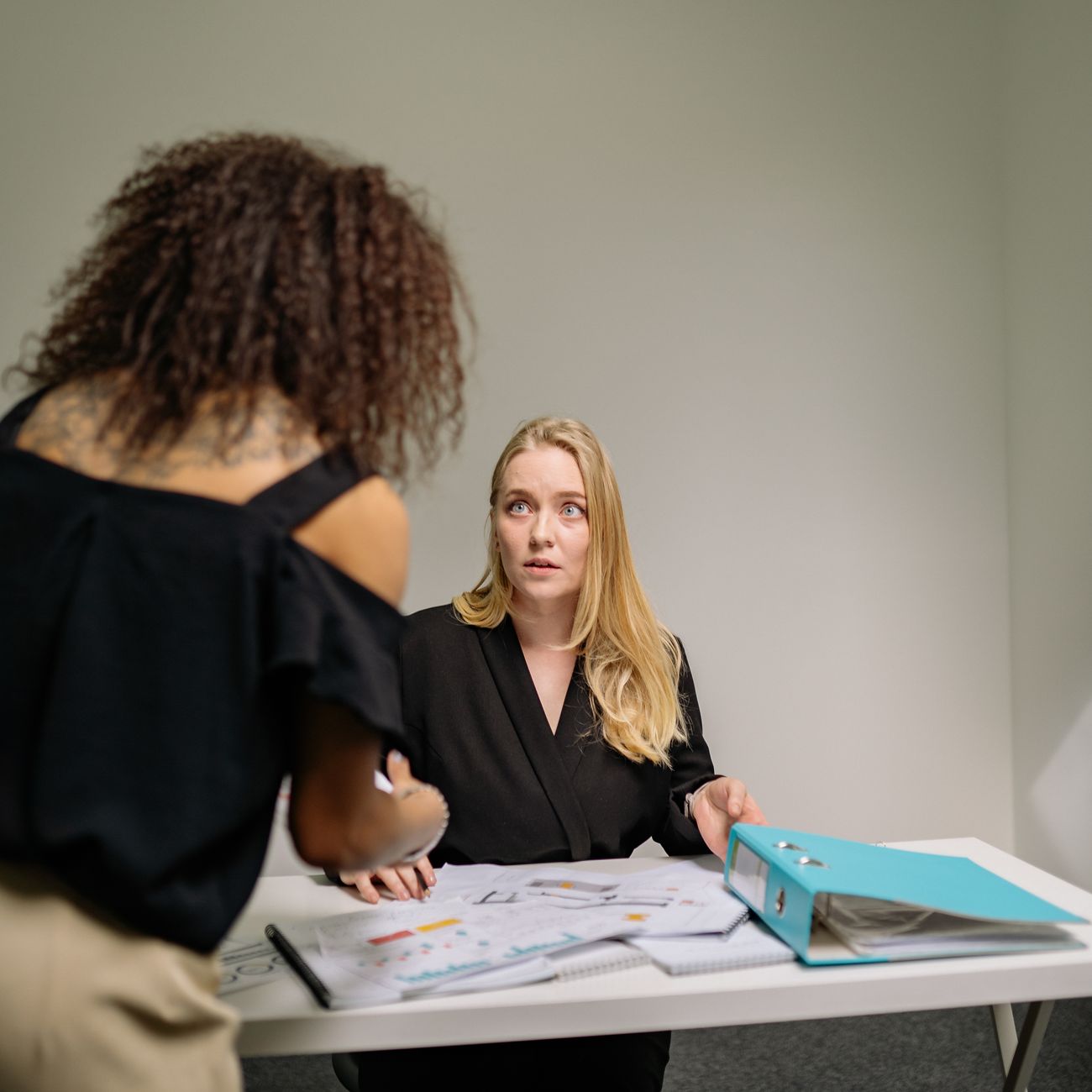 Two women sitting at an office desk reviewing documents and a binder together
