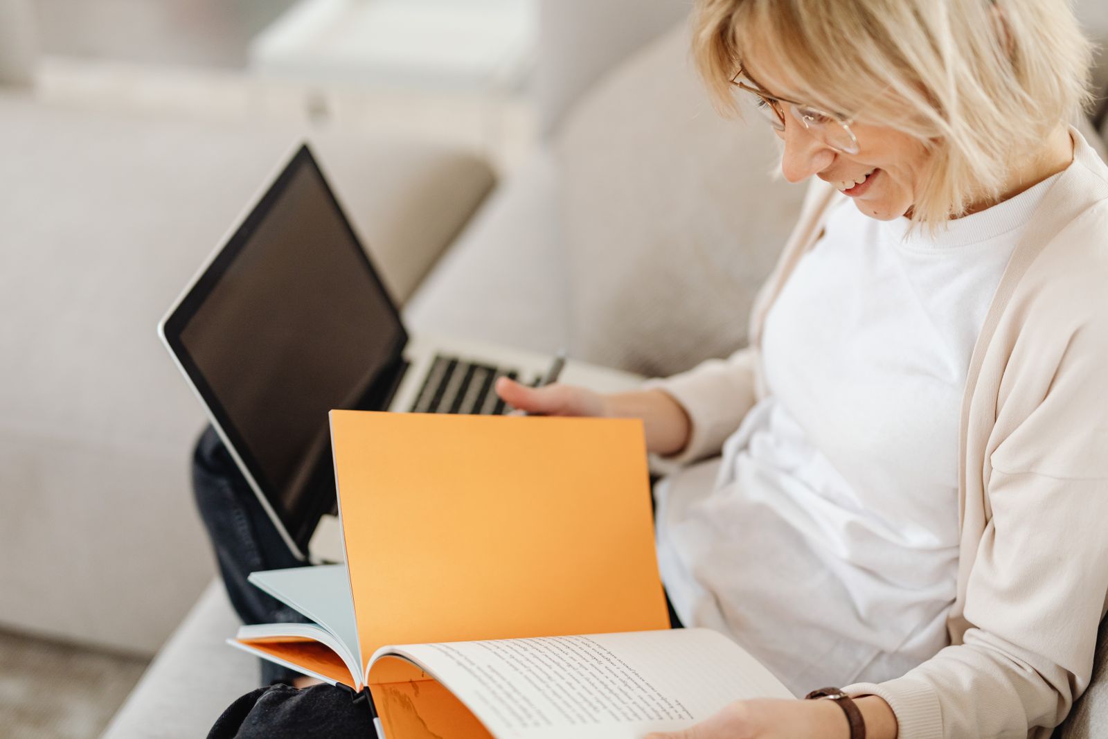 A woman in glasses sitting on a sofa, reading an open book with a pen in hand and a laptop on her lap